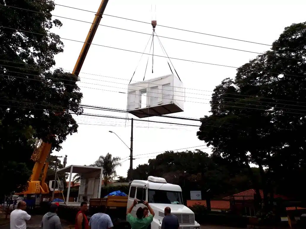 Instalação de um elemento pré-fabricado com guindaste por uma equipe de trabalhadores, com uma caminhonete ao fundo, árvores e fios de energia ao redor.